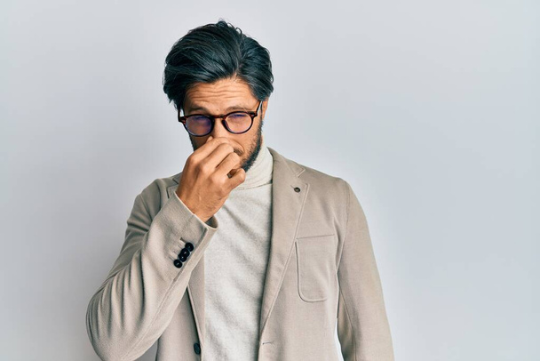 Young hispanic man wearing business jacket and glasses smelling something stinky and disgusting, intolerable smell, holding breath with fingers on nose. bad smell  - Fotoğraf, Görsel
