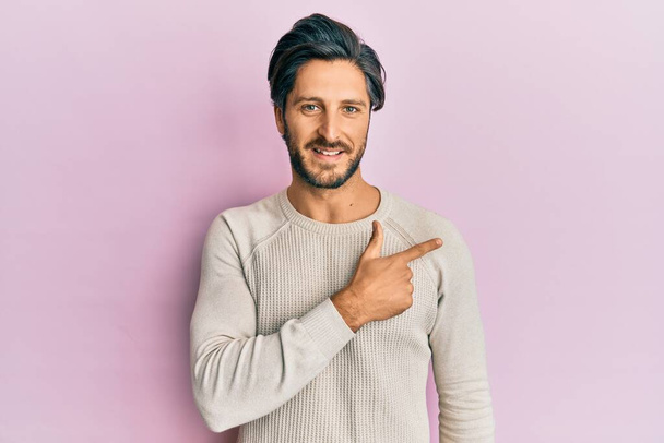 Fiatal spanyol férfi alkalmi téli pulóvert visel vidám mosollyal az arcán, kezével és ujjával oldalra mutat, boldog és természetes arckifejezéssel.  - Fotó, kép