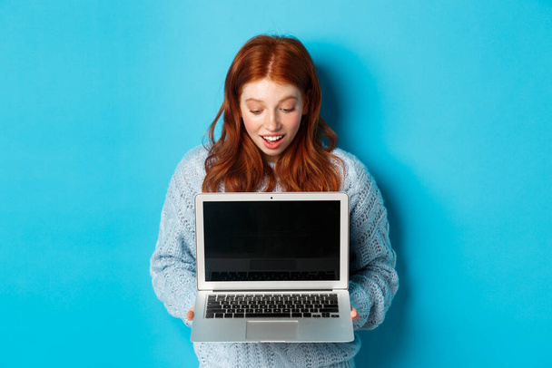 Úžasný ryšavý dívka zírá na obrazovce notebooku a vypadá ohromeně, ukazuje počítačový displej, stojí nad modrým pozadím - Fotografie, Obrázek