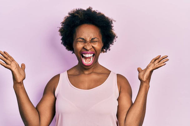 Νεαρή Αφροαμερικάνα που φοράει casual αμάνικο t-shirt γιορτάζοντας τρελή και τρελή για την επιτυχία με τα χέρια σηκωμένα και τα κλειστά μάτια να ουρλιάζουν ενθουσιασμένα. έννοια νικητή  - Φωτογραφία, εικόνα