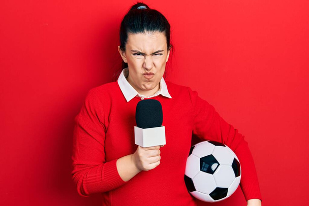 Молодая латиноамериканка держит в руках репортера микрофон и футбольный мяч скептически и нервно, хмурясь расстроена из-за проблем. отрицательный человек.  - Фото, изображение