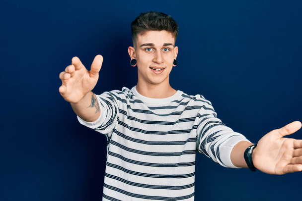 Young caucasian boy with ears dilation wearing casual striped shirt looking at the camera smiling with open arms for hug. cheerful expression embracing happiness.  - Photo, Image