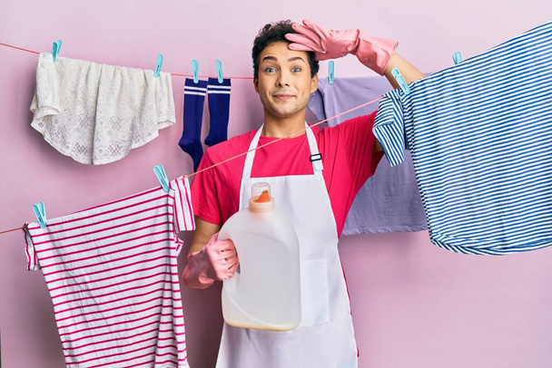 Hombre hispano guapo lavando la ropa sosteniendo botella de detergente estresado y frustrado con la mano en la cabeza, cara sorprendida y enojada  - Foto, Imagen