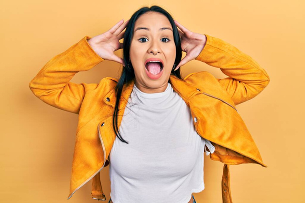 Beautiful hispanic woman with nose piercing wearing yellow leather jacket crazy and scared with hands on head, afraid and surprised of shock with open mouth  - Photo, Image