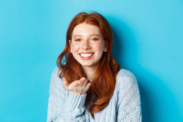 Mooie roodharige vrouw model glimlachen, het verzenden van lucht kus op de camera, staande tegen een blauwe achtergrond - Foto, afbeelding