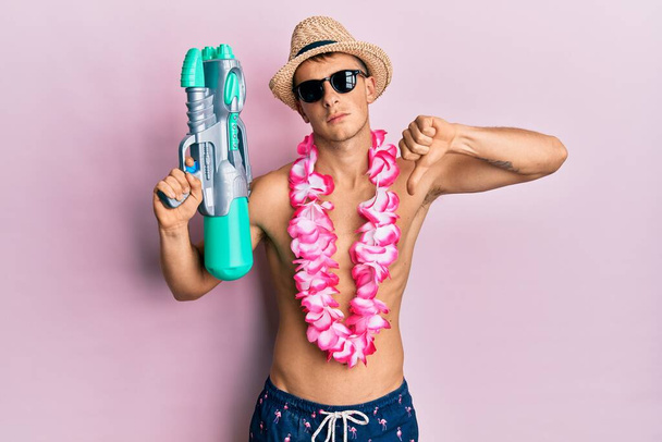 Young caucasian man wearing swimsuit and hawaiian lei holding watergun with angry face, negative sign showing dislike with thumbs down, rejection concept  - Photo, Image