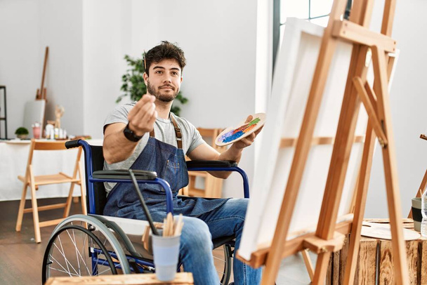 Young hispanic man sitting on wheelchair painting at art studio doing money gesture with hands, asking for salary payment, millionaire business  - Photo, image