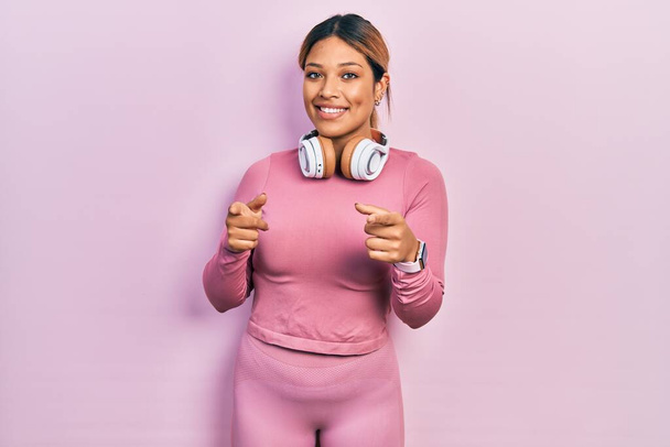 Beautiful hispanic woman wearing gym clothes and using headphones pointing fingers to camera with happy and funny face. good energy and vibes.  - Photo, Image