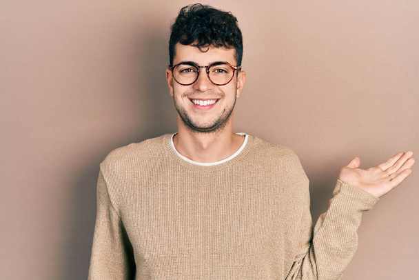 Young hispanic man wearing casual clothes and glasses smiling cheerful presenting and pointing with palm of hand looking at the camera.  - Foto, imagen
