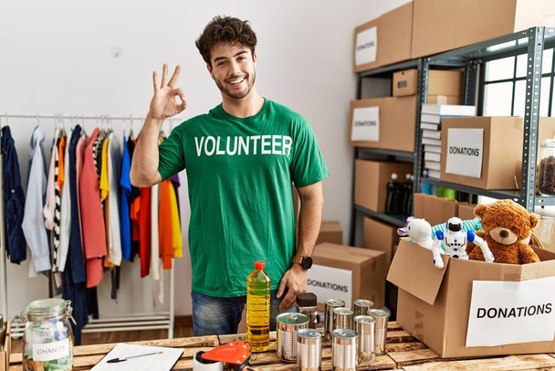 Bağışta gönüllü tişört giyen İspanyol adam tamam işareti yapıyor, gülümsüyor ve mükemmel bir sembol oluşturuyor.  - Fotoğraf, Görsel