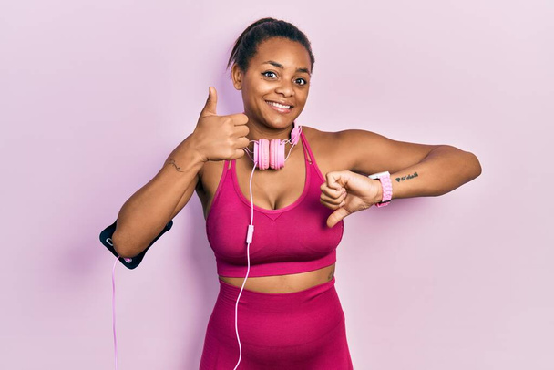 Jong Afrikaans Amerikaans meisje draagt sportkleding en gebruikt koptelefoons die duimen op en neer doen, onenigheid en eensgezindheid. gek conflict  - Foto, afbeelding