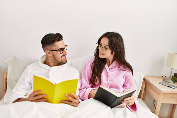 Joven pareja hispana leyendo libro acostada en la cama en casa. - Foto, imagen