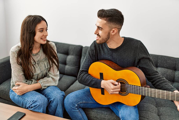 Молодая испаноязычная пара, играющая на испанской гитаре, сидит дома на диване. - Фото, изображение
