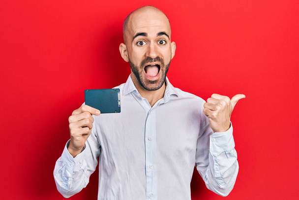 Young bald man holding ssd memory pointing thumb up to the side smiling happy with open mouth  - Photo, image
