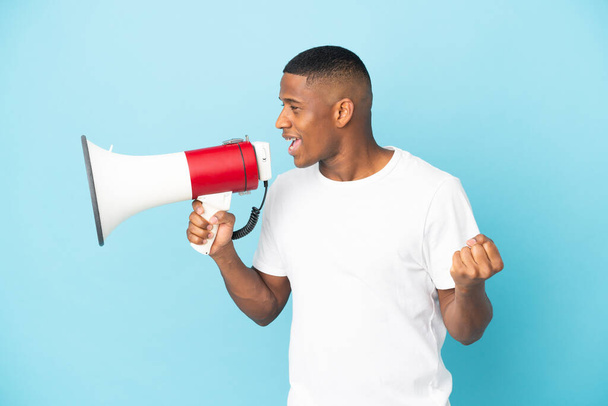 Young latin man isolated on blue background shouting through a megaphone to announce something in lateral position - Photo, Image