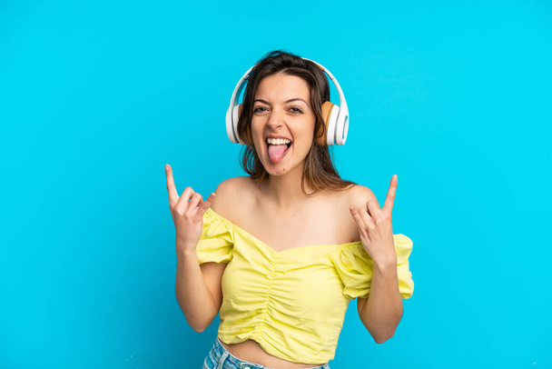 Молодая белая женщина, изолированная на синем фоне, слушает музыку, делая рок-жест - Фото, изображение