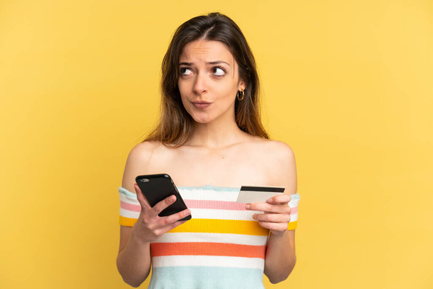 Νεαρή καυκάσια γυναίκα απομονωμένη σε κίτρινο φόντο αγοράζει με το κινητό με πιστωτική κάρτα, ενώ σκέφτεται - Φωτογραφία, εικόνα