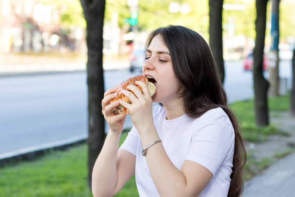 Mulher morena comendo hambúrguer na rua perto da estrada na cidade. Fast food a caminho do trabalho ou da escola. - Foto, Imagem