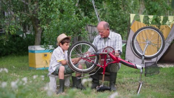 rodičovství, starostlivý starší muž s chlapcem v klobouku bavit a opravit kolo při sezení na zahradě v létě - Záběry, video