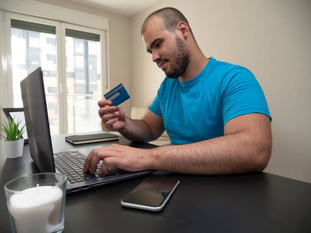 Egy fiatalember dolgozik a nappalijából egy laptopon egy fekete asztalon egy fehér gyertyával a telefonjával és egy hitelkártyával egy fekete noteszral. - Fotó, kép