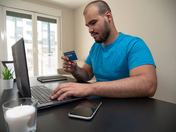 Νεαρός που εργάζεται από το σαλόνι του σε ένα φορητό υπολογιστή σε ένα μαύρο τραπέζι με ένα λευκό κερί το τηλέφωνό του και μια πιστωτική κάρτα με ένα μαύρο σημειωματάριο - Φωτογραφία, εικόνα