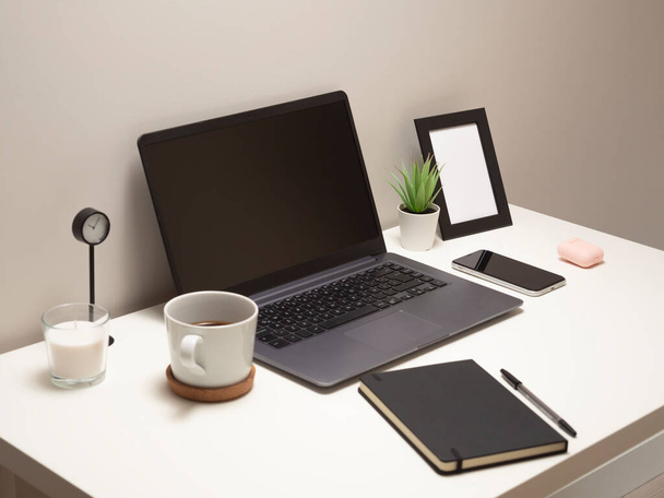 Bild vom Laptop auf einem weißen Tisch mit einem Becher eine Uhr ein kleiner Busch ein Notizbuch ein Telefon ein Bilderrahmen und eine Kopfhörertasche - Foto, Bild