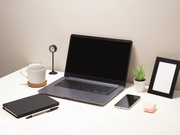 Foto van een laptop op een witte tafel met een mok een klok een kleine struik een notebook een telefoon een fotolijstje en een koptelefoonhoes - Foto, afbeelding