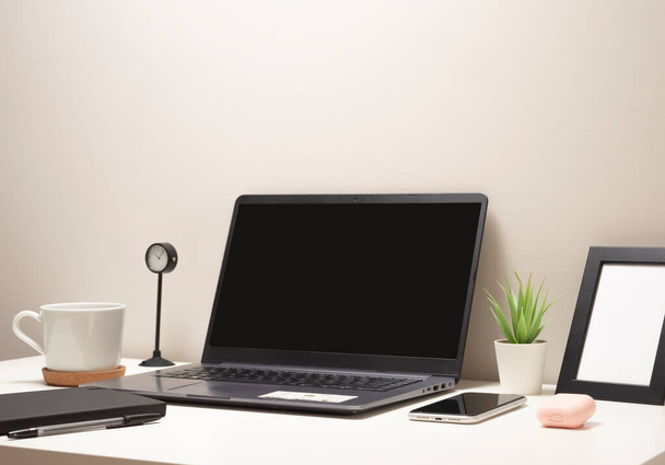 Imagem de computador portátil em uma mesa branca com uma caneca um relógio um pequeno arbusto um caderno um telefone uma armação de imagem e um caso de fone de ouvido - Foto, Imagem