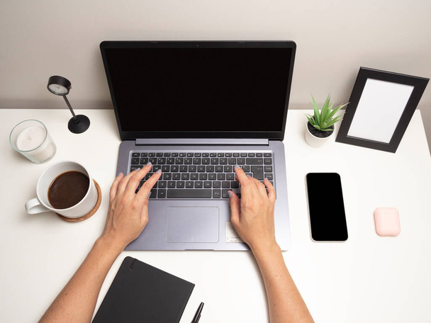 Ręce kobiety na białym stole z laptopem wpisującym się na klawiaturze obok telefonu komórkowego czarny notatnik filiżanka kawy zegar biała świeca mały krzak i pusta ramka na zdjęcia - Zdjęcie, obraz