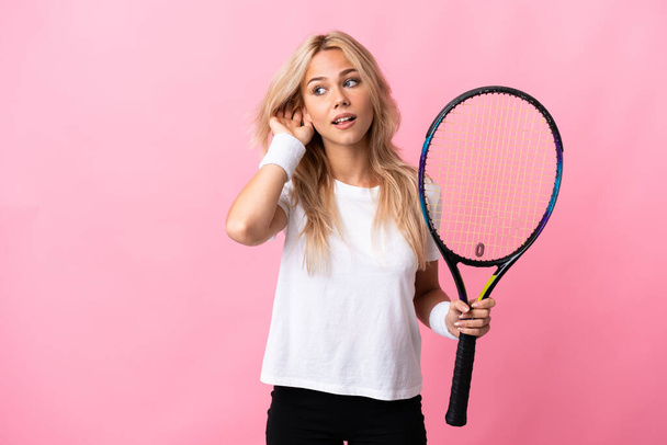 junge russische Frau spielt Tennis isoliert auf violettem Hintergrund und hört etwas, indem sie die Hand auf das Ohr legt - Foto, Bild