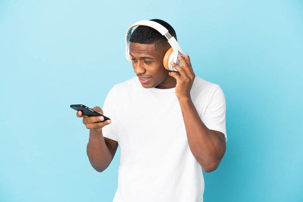 młody Latynos człowiek odizolowany na niebieskim tle słuchanie muzyki z telefonu komórkowego i śpiew - Zdjęcie, obraz