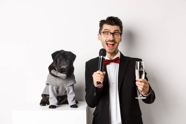 Feliz joven dando un discurso y levantando champán de cristal, de pie con lindo perro negro en traje de fiesta sobre fondo blanco - Foto, Imagen