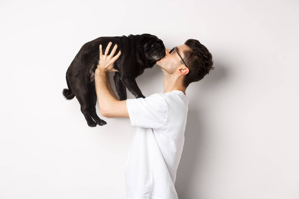 Profile of handsome young man kissing small cute dog face. Hipster guy loving his pug, standing over white background - Zdjęcie, obraz