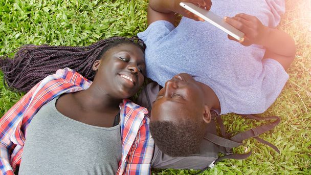 Αφρικανό ζευγάρι εφήβων ξαπλωμένο στο γρασίδι χρησιμοποιώντας tablet και χαλαρωτικό. Έννοια ευτυχίας και ελαφρότητας. - Φωτογραφία, εικόνα