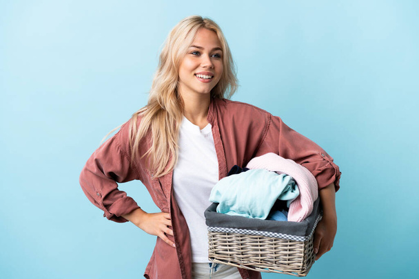 Elinde mavi arka planda izole edilmiş bir çamaşır sepeti tutan genç Rus kadın kolları kalçasında ve gülümsüyor. - Fotoğraf, Görsel
