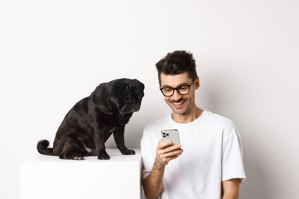 Joven sonriente usando smartphone y sentado cerca del perro. Propietario Pug comprobar fotos en el teléfono móvil, fondo blanco - Foto, Imagen