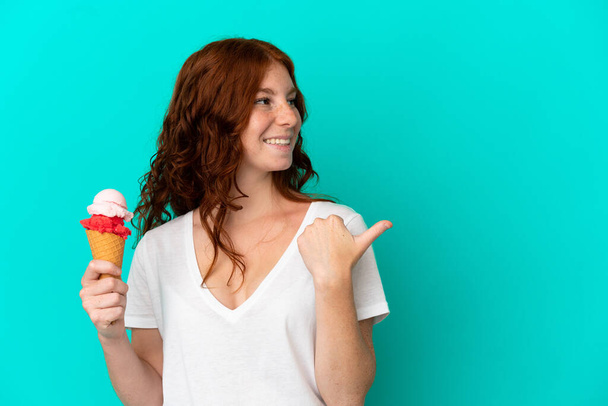 Teenager reddish woman with a cornet ice cream isolated on blue background pointing to the side to present a product - Fotografie, Obrázek