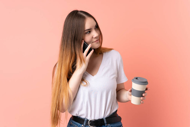 Young caucasian woman isolated on pink background holding coffee to take away and a mobile - Photo, Image