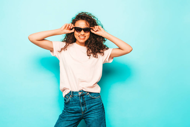 Beautiful black woman with afro curls hairstyle.Smiling model in white trendy t-shirt and jeans clothes. Sexy carefree female posing near blue in studio. Tanned and cheerful. In sunglasses - Fotoğraf, Görsel