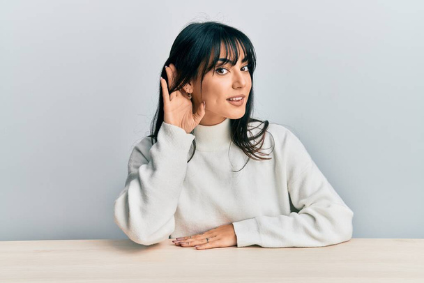 Young brunette woman with bangs wearing casual clothes sitting on the table smiling with hand over ear listening and hearing to rumor or gossip. deafness concept.  - Photo, Image