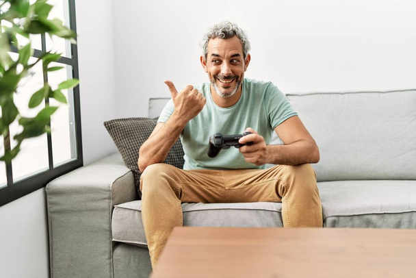 Moyen Age hispanique homme jouant jeu vidéo assis sur le canapé pointant pouce vers le côté souriant heureux avec la bouche ouverte  - Photo, image