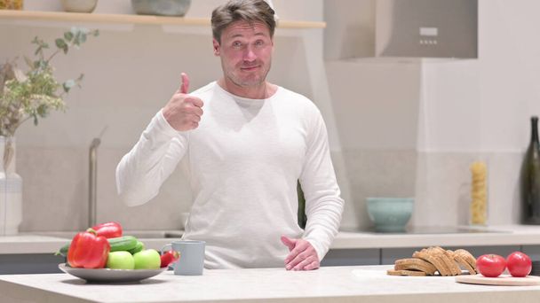 Чоловік середнього віку показує великі пальці вгору, стоячи на кухні
 - Фото, зображення