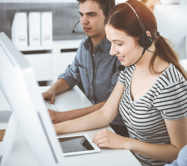 Casual dressed young woman using headset and computer while talking with customers online. Group of operators at work in sunny office. Call center - 写真・画像