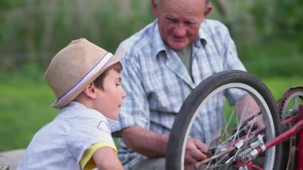 niño varón feliz con el abuelo se regocija en la reparación de la rueda en bicicleta y levanta las manos haciendo vítores gesto telón de fondo de árboles verdes - Imágenes, Vídeo