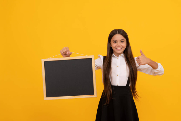 χαρούμενο έφηβο κορίτσι με στολή κρατήστε τον πίνακα του σχολείου για να εμφανιστεί ο αντίχειρας, παρουσιάζοντας - Φωτογραφία, εικόνα