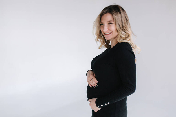 Mulher grávida elegante em mãos vestido preto de inicialização na barriga e sorrir. Fundo branco - Foto, Imagem