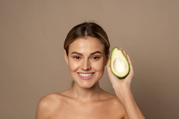 Šťastně se usmívající mladá žena drží avokádo napůl na béžovém pozadí. Koncept přírodní krásy - Fotografie, Obrázek