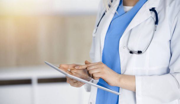 Unknown woman-doctor standing in sunny clinic and using tablet pc, close-up. Data and best service in medicine - Photo, image