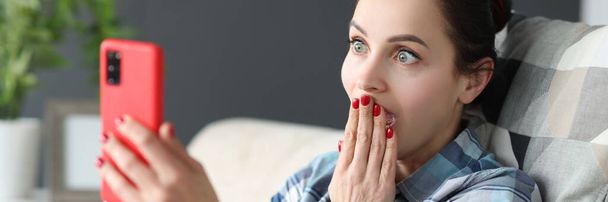 Surprised woman looks at smartphone screen while sitting on couch - Фото, изображение