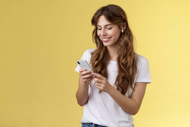 Happy caring tender feminine girlfriend curly long hair hold smartphone picking song listen way home smiling broadly look mobile phone scroll music platform wear wireless earbud yellow background - Photo, Image